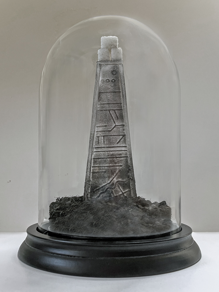 a glass dome protects a sculpture that's the shape of a tall plinth, tapered at the top. On top are 3 sugar cubes. At the base is a layer of black peat.