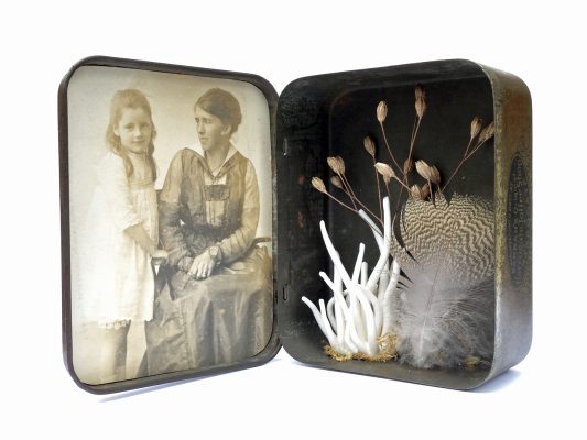 an old tin sits open. Inside the lid is a photograph of a Victorian woman looking at a child. Inside the tin is a feather, a dried seedhead and a white 'lichen' made of porcelain