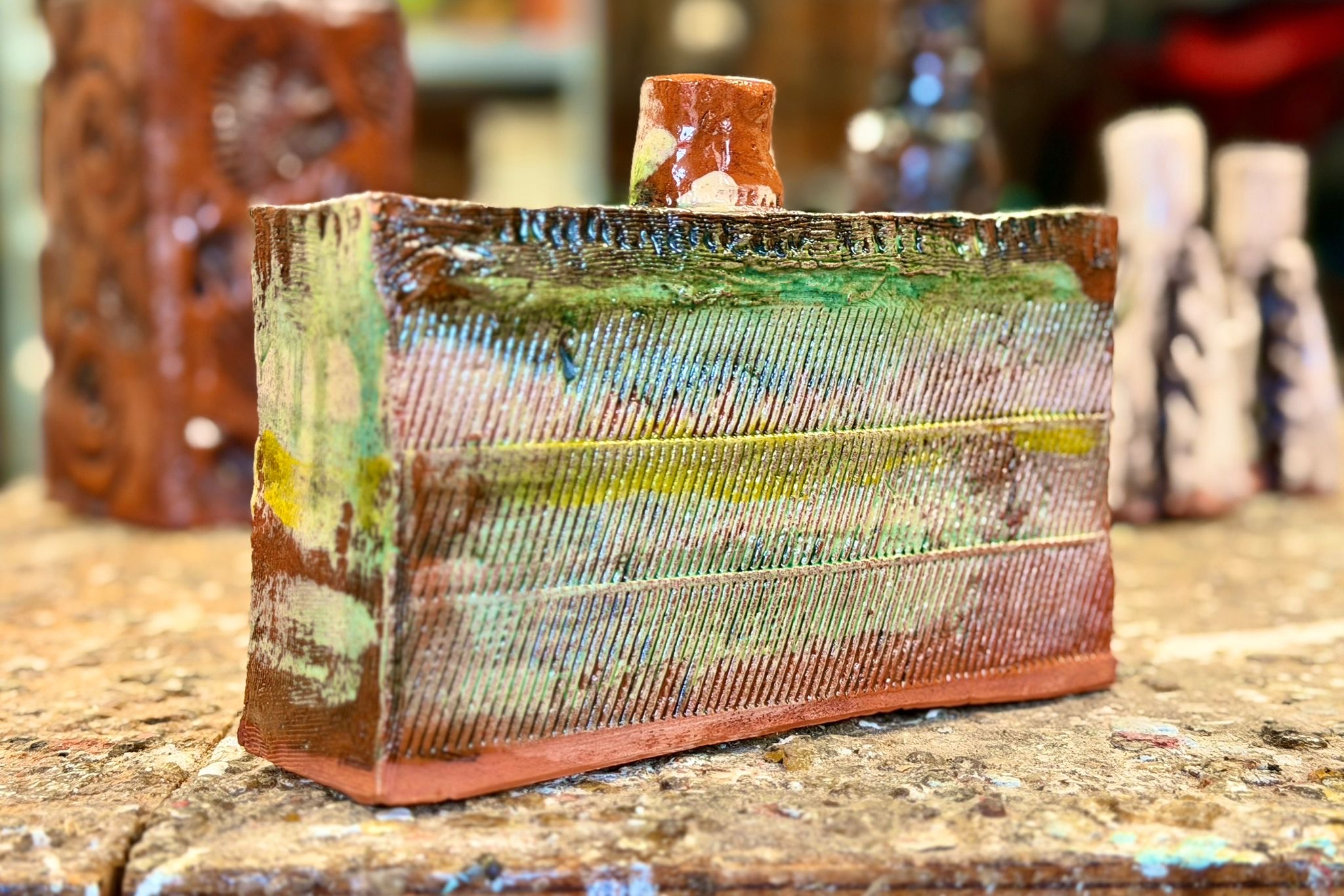 Image of pottery bottle. It is rectangular and is coloured with brown and green, sat on a table