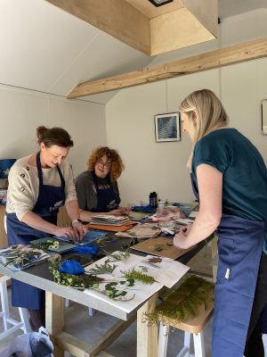 3 ladies smiling while placing their foraged plants onto the sensitised paper to make cyanotype cards at a workshop.